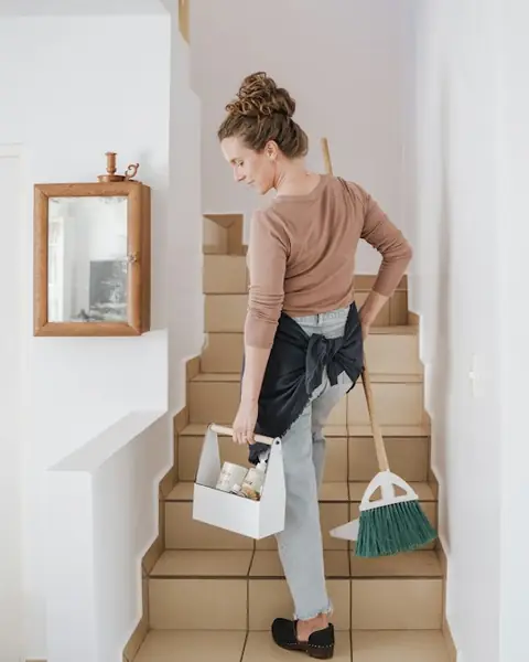 Someone carrying a cleaning caddy and broom up a flight of stairs at home.