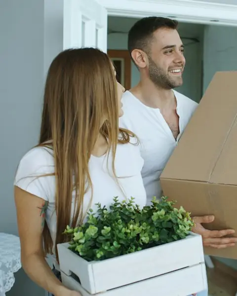 Two people carrying moving boxes and a plant into a new home.